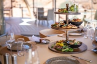 A beautifully set dining table with a three-tiered serving tray filled with a variety of gourmet appetizers. Fresh vegetables, bread, and small dishes of food are arranged artistically. The table is adorned with elegant glassware and an assortment of plates and cutlery, set in a bright, sunlit setting, suggesting a luxurious dining experience.