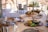 A beautifully set dining table with a three-tiered serving tray filled with a variety of gourmet appetizers. Fresh vegetables, bread, and small dishes of food are arranged artistically. The table is adorned with elegant glassware and an assortment of plates and cutlery, set in a bright, sunlit setting, suggesting a luxurious dining experience.