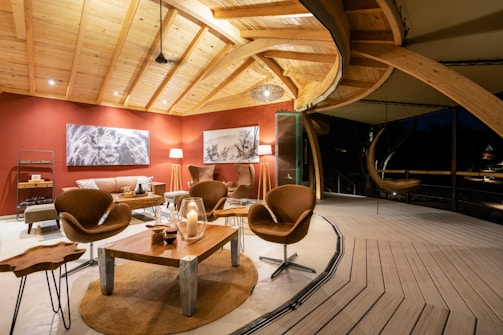 A luxurious, modern living area with a high wooden ceiling and warm lighting. The room features stylish brown leather chairs, a wooden coffee table with decorative items, and a few pieces of contemporary art on the red walls. There's an open, airy feel with large glass windows allowing views of the outside. Subtle lighting creates an inviting and cozy atmosphere.