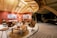 A luxurious, modern living area with a high wooden ceiling and warm lighting. The room features stylish brown leather chairs, a wooden coffee table with decorative items, and a few pieces of contemporary art on the red walls. There's an open, airy feel with large glass windows allowing views of the outside. Subtle lighting creates an inviting and cozy atmosphere.