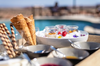 A wooden tray holds a variety of desserts in small glass jars, each topped with colorful fruits and edible flowers. Next to the jars, several empty waffle cones are placed in a glass. Striped wafer sticks are also visible. The setting is outdoors with a pool in the blurred background.