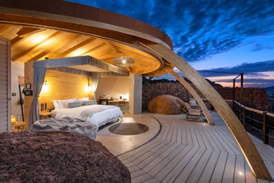 A luxurious outdoor bedroom features a modern canopy bed with white bedding and decorative pillows. Wooden beams create a warm and inviting roof structure above. The space is illuminated by soft lighting, accentuating the natural wood textures. A circular rug lies on the deck, which extends to an open view of the twilight sky. Large rocks and rustic stone walls enhance the earthy ambiance. Several chairs are arranged on the deck area for lounging.