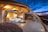 A luxurious outdoor bedroom features a modern canopy bed with white bedding and decorative pillows. Wooden beams create a warm and inviting roof structure above. The space is illuminated by soft lighting, accentuating the natural wood textures. A circular rug lies on the deck, which extends to an open view of the twilight sky. Large rocks and rustic stone walls enhance the earthy ambiance. Several chairs are arranged on the deck area for lounging.