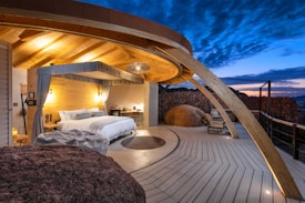 A luxurious outdoor bedroom features a modern canopy bed with white bedding and decorative pillows. Wooden beams create a warm and inviting roof structure above. The space is illuminated by soft lighting, accentuating the natural wood textures. A circular rug lies on the deck, which extends to an open view of the twilight sky. Large rocks and rustic stone walls enhance the earthy ambiance. Several chairs are arranged on the deck area for lounging.
