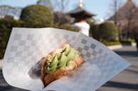 A dessert consisting of a pastry filled with green matcha ice cream is held in a checkered paper wrapper. In the background, there is a garden setting with shrubs and a blurred structure resembling an Asian-style roof.
