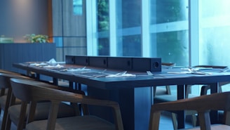 A modern dining room setup features a long wooden table set with cutlery and napkins, surrounded by wooden chairs. Large floor-to-ceiling windows allow natural light to fill the space, and a plant adds a decorative touch. The overall design is sleek and contemporary.