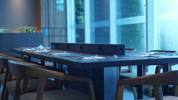 A modern dining room setup features a long wooden table set with cutlery and napkins, surrounded by wooden chairs. Large floor-to-ceiling windows allow natural light to fill the space, and a plant adds a decorative touch. The overall design is sleek and contemporary.