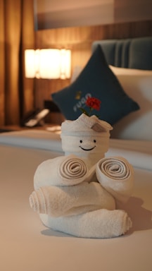 A creative towel sculpture resembling a figure with a smiling face, adorned with a small red flower on top. It is placed on a neatly made bed in a cozy and warmly lit room. In the background, there is a teal pillow and a bedside light next to a telephone.