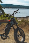 A rugged electric bike is positioned on a grassy area near a scenic body of water, surrounded by lush greenery and distant hills under a clear sky.