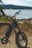 A rugged electric bike is positioned on a grassy area near a scenic body of water, surrounded by lush greenery and distant hills under a clear sky.