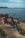 A dirt path leads to a picturesque view of the ocean with a blue electric bike parked next to a pink wooden chair. The scene is set against a backdrop of a calm, expansive body of water stretching to the horizon with a strip of land in the distance.