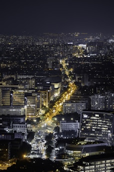 A vibrant cityscape at night with numerous illuminated buildings and a prominent street lined with bright lights. The density of the buildings and the glow of the city lights create an intricate and lively urban scene.