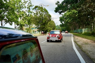 A classic red car with white racing stripes drives along a tree-lined road. The car is followed by several other vehicles, and the road is bordered by lush greenery on both sides. The sky above is partly cloudy, creating a serene and peaceful driving environment.
