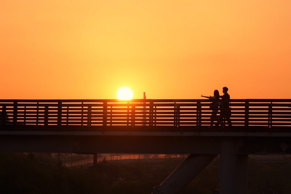 a couple of people walking across a bridge at sunset
