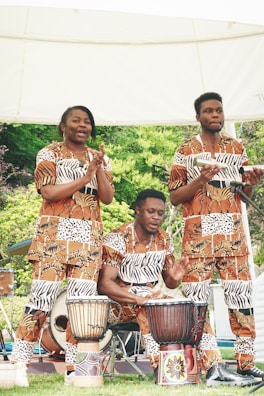 Three individuals dressed in matching animal print outfits are performing music outdoors. Two of them are playing African drums, while the third claps along. The setting appears to be in a park or garden area with lush greenery in the background.