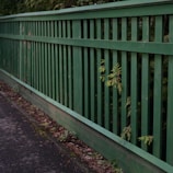A wooden fence painted in green runs alongside a paved pathway. The vertical slats of the fence allow some greenery to poke through, adding a natural touch to the man-made structure. The pathway appears slightly worn with scattered leaves and debris.