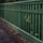 A wooden fence painted in green runs alongside a paved pathway. The vertical slats of the fence allow some greenery to poke through, adding a natural touch to the man-made structure. The pathway appears slightly worn with scattered leaves and debris.