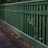 A wooden fence painted in green runs alongside a paved pathway. The vertical slats of the fence allow some greenery to poke through, adding a natural touch to the man-made structure. The pathway appears slightly worn with scattered leaves and debris.