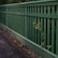 A wooden fence painted in green runs alongside a paved pathway. The vertical slats of the fence allow some greenery to poke through, adding a natural touch to the man-made structure. The pathway appears slightly worn with scattered leaves and debris.