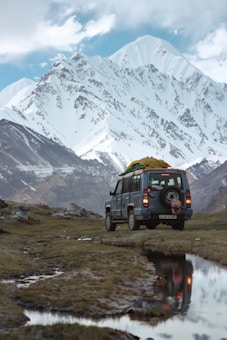 An off-road vehicle is parked on a grassy terrain with snow-capped mountains towering in the background. The vehicle has luggage secured on its roof, and a reflection can be seen in a small puddle nearby. The scene is set against a partly cloudy sky, creating a picturesque landscape.