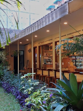 An exterior view of a cozy cafe with an outdoor seating area. The cafe has a modern design with large windows, warm lighting from the ceiling, and wooden bar stools lined up along a counter. A row of lush green plants and colorful flowers lines the pathway leading to the entrance, enhancing the tranquil ambiance. Above the seating area, a canopy displays text.