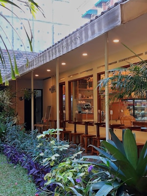 An exterior view of a cozy cafe with an outdoor seating area. The cafe has a modern design with large windows, warm lighting from the ceiling, and wooden bar stools lined up along a counter. A row of lush green plants and colorful flowers lines the pathway leading to the entrance, enhancing the tranquil ambiance. Above the seating area, a canopy displays text.