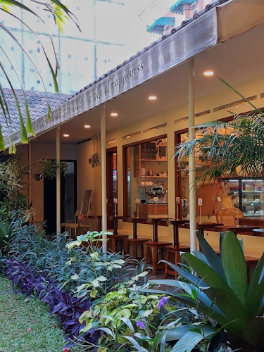 An exterior view of a cozy cafe with an outdoor seating area. The cafe has a modern design with large windows, warm lighting from the ceiling, and wooden bar stools lined up along a counter. A row of lush green plants and colorful flowers lines the pathway leading to the entrance, enhancing the tranquil ambiance. Above the seating area, a canopy displays text.
