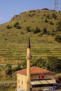 A tall minaret attached to a stone building with a red tiled roof, displaying a Turkish flag. The structure is set against a backdrop of a green hillside with scattered trees. There are communication towers at the top of the hill under a clear blue sky.