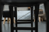 A unique perspective showing the underside of a chair, revealing a modern, minimalist interior with two stools featuring wooden legs. The background includes a textured concrete wall and additional woven furniture pieces, creating an industrial yet cozy atmosphere.