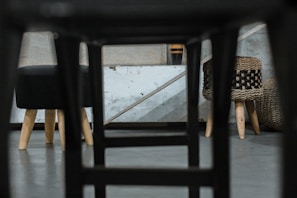 A unique perspective showing the underside of a chair, revealing a modern, minimalist interior with two stools featuring wooden legs. The background includes a textured concrete wall and additional woven furniture pieces, creating an industrial yet cozy atmosphere.