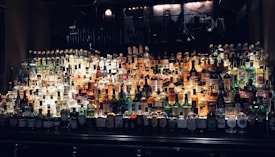 A well-stocked bar filled with an extensive variety of liquor bottles arranged on shelves. Bottles of different shapes, sizes, and colors are displayed, including well-known brands and diverse types of spirits. The backlighting highlights the bottles, giving them a warm glow and making the labels visible.