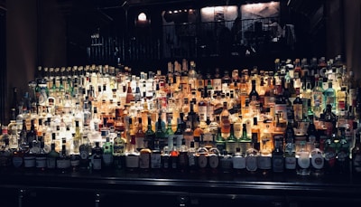 A well-stocked bar filled with an extensive variety of liquor bottles arranged on shelves. Bottles of different shapes, sizes, and colors are displayed, including well-known brands and diverse types of spirits. The backlighting highlights the bottles, giving them a warm glow and making the labels visible.