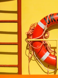 A bright yellow surface with a vivid red life preserver ring attached to it. The life preserver has white accents and is secured with coiled orange rope. To the left, part of a red ladder-like structure is visible.