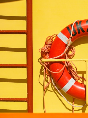 A bright yellow surface with a vivid red life preserver ring attached to it. The life preserver has white accents and is secured with coiled orange rope. To the left, part of a red ladder-like structure is visible.