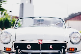 A close-up view of the front of a white vintage MG convertible car, featuring the iconic MG badge on the grill. The car is parked outdoors, likely in a sunny area as indicated by the bright lighting and shadows. The background includes some greenery and the blurred outline of buildings.