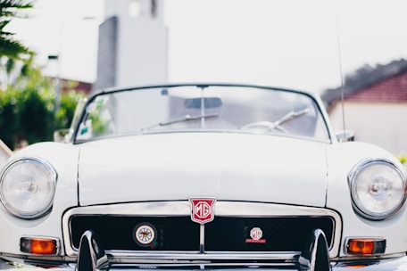 A close-up view of the front of a white vintage MG convertible car, featuring the iconic MG badge on the grill. The car is parked outdoors, likely in a sunny area as indicated by the bright lighting and shadows. The background includes some greenery and the blurred outline of buildings.