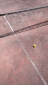 A tennis court with a net stretching across the middle and a yellow tennis ball resting near the net on the clay surface. The court lines are faintly visible, and the ground appears to have a reddish-brown tone.