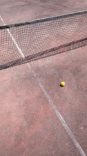 A tennis court with a net stretching across the middle and a yellow tennis ball resting near the net on the clay surface. The court lines are faintly visible, and the ground appears to have a reddish-brown tone.