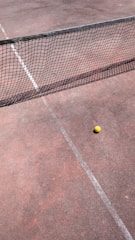 A tennis court with a net stretching across the middle and a yellow tennis ball resting near the net on the clay surface. The court lines are faintly visible, and the ground appears to have a reddish-brown tone.