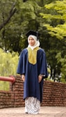 A person wearing a graduation cap and gown stands on a brick pathway lined with a red railing. The background is a lush green setting with trees and foliage.