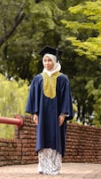 A person wearing a graduation cap and gown stands on a brick pathway lined with a red railing. The background is a lush green setting with trees and foliage.