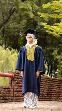 A person wearing a graduation cap and gown stands on a brick pathway lined with a red railing. The background is a lush green setting with trees and foliage.
