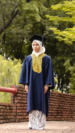 A person wearing a graduation cap and gown stands on a brick pathway lined with a red railing. The background is a lush green setting with trees and foliage.