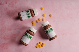 Three brown pill bottles with white caps, each labeled with green and white text, are scattered on a pink textured surface. Between the bottles, several small, round, yellow tablets are arranged in clusters.