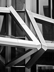 A complex architectural facade composed of intersecting metal beams, creating an intricate geometric pattern. The reflective surfaces of the beams and the surrounding glass windows play with light and shadow, adding depth and contrast. The design appears modern and futuristic.