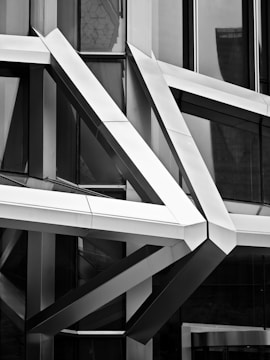 A complex architectural facade composed of intersecting metal beams, creating an intricate geometric pattern. The reflective surfaces of the beams and the surrounding glass windows play with light and shadow, adding depth and contrast. The design appears modern and futuristic.