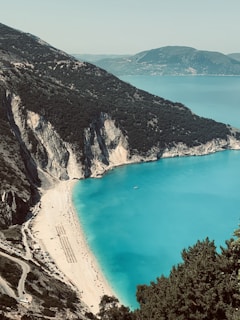 A picturesque coastal landscape featuring a curved sandy beach bordered by steep, rocky cliffs. The turquoise sea contrasts with the dark green foliage on the cliffs. Below, rows of sunbeds and umbrellas are lined up, indicating a popular beach destination. In the distance, rolling hills meet the horizon under a clear sky.