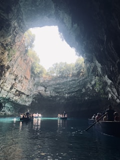 A stunning cave with a large open ceiling allowing natural light to illuminate the interior. The clear blue water reflects the light, and several small boats with people are floating on the water. Lush greenery can be seen growing around the upper edges of the cave.