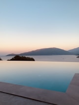 An infinity pool with a view of calm waters extending to the horizon, bordered by mountains on either side. The sky is a picturesque gradient transitioning from warm hues of orange near the horizon to a soft blue overhead, indicating either sunrise or sunset.