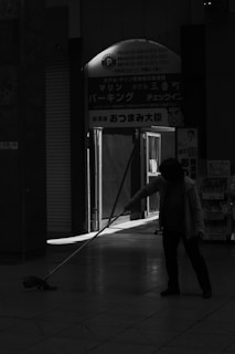 A person is mopping the floor inside a dimly lit interior space with a bright doorway in the background. The image is in black and white, creating a high-contrast effect with shadows and light.